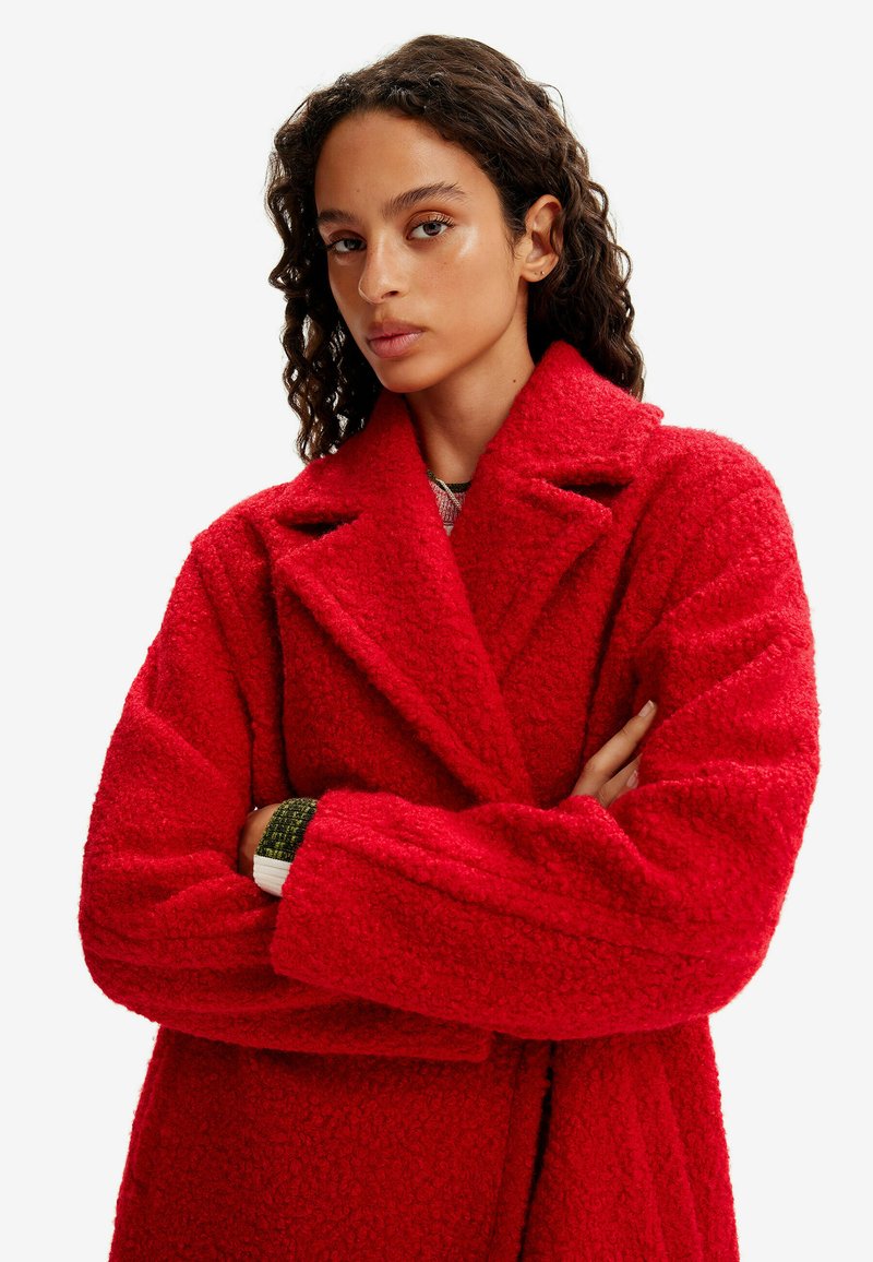 Donna con capelli ricci scuri che indossa un cappotto rosso brillante e strutturato, con le braccia incrociate, che guarda in avanti su uno sfondo bianco uniforme.