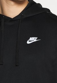 Schwarze Kapuzenjacke aus weichem Material mit einem zentralen weißen Nike-Logo, ausgestattet mit einer Kordelzugkapuze und strukturierten Schulterakzenten.