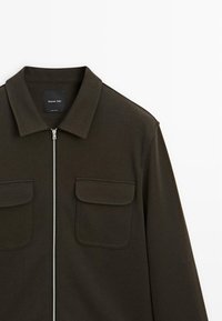 Mørkeolivengr�øn zip-up jakke med krave, to brystlommer og et struktureret stof. Brandingmærke synligt ved kraven.