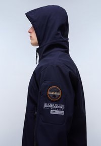 Osoba w granatowej kurtce z kapturem z naszywką z logo Napapijri i napisem na górnej części ramienia, zwrócona w lewo na jednolitym jasnym tle.