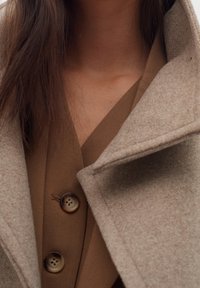 Cappotto in misto lana beige con ampio colletto, dotato di due grandi bottoni rotondi scuri, indossato sopra un top marrone con scollatura a V e tessuto liscio.