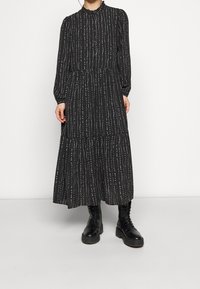 Dlouhá černá šaty s bílými svislými pruhy, s vysokým límcem, zapínáním na knoflíky vpředu, dlouhými rukávy a volánovou sukní.