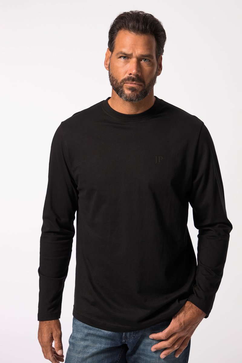 Hombre con barba, vestido con camisa negra de manga larga y vaqueros azules, de pie frente a un fondo liso y claro con expresión seria.