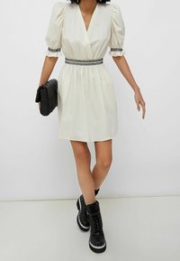 Hvid kjole med pufærmer, V-udskæring og sort broderet talje, parret med en sort vævet clutch og sorte kampstøvler.