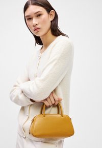 Geel leren handtas met een gestructureerde, ronde vorm, dubbele handvatten aan de bovenkant en ritssluiting. Draagt een model in een lichte, textuurcardigan.