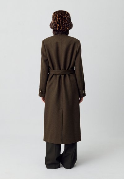 Cappotto lungo in lana di un verde oliva, con vita cintrata e maniche a punca, indossato sopra pantaloni neri a gamba larga. Accessoriato con un cappello a secchiello in stampa leopardata.