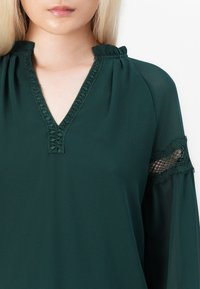 Grønn, flytende bluse med V-hals og dekorativ søm, utsvingte ermer med blonder, og samling detaljer ved skuldrene.