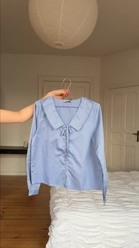 Hellblaue Bluse mit langen Ärmeln, gewelltem Kragen und vorderen Knöpfen, gehalten an einem Bügel in einem Schlafzimmer.