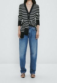 Czarnobiały sweter w paski z wiązaniem w talii, z głębokim dekoltem w serek, dwoma guzikami, noszony na luźne niebieskie dżinsy i czarne sandały z paskami.