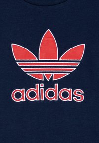 Tricou din bumbac de un albastru marin, având un logo brodat Adidas în culorile roșu și alb, reprezentând un trifoi cu trei frunze și denumirea brandului dedesubt.