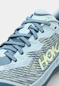 Light blue athletic shoe features a textured upper, grey laces, and a patterned side with a prominent yellow brand logo.