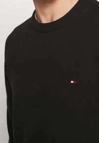 Pull en maille noire avec un col côtelé. Présente un petit logo brodé avec des accents rouges, blancs et bleus sur la poitrine gauche.