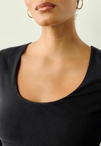 Melna topiņa ar lašu kakla izgriezumu, izgatavota no mīksta, elastīga auduma; gluda tekstūra ar nelielu spīdumu; tuvplāns uz augšējo krūtīm un kakla izgriezumu.