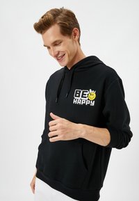 Sudadera negra de tejido suave, con un bolsillo canguro y un gráfico blanco que dice "BE HAPPY" acompañado de una carita sonriente amarilla y acentos.