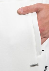 Pantalones de chándal blancos con una textura suave, que cuentan con un bolsillo lateral y una etiqueta de metal etiquetada "SMILODOX" cerca del dobladillo.