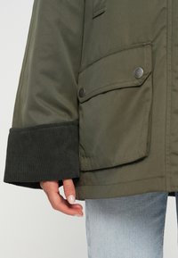 Κοντινό πλάνο τσέπης πράσινου μπουφάν με κουμπιά πατιλέτας, ribbed μαύρη μανσέτα, φορεμένο με ανοιχτό μπλε τζιν και ορατό χέρι στο πλάι.