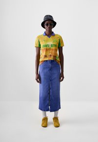 Maglia da calcio gialla e verde con testo grafico, abbinata a una lunga gonna di jeans. Indossata con scarpe da ginnastica gialle e calzini bianchi. Cappello a secchiello nero.