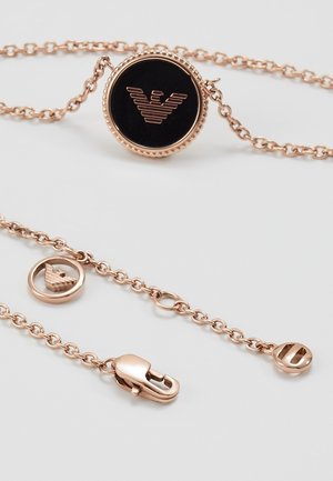 Collier chaîne en or rose avec pendentif noir circulaire arborant un logo d'aigle et un fermoir mousquetaire sur fond blanc.