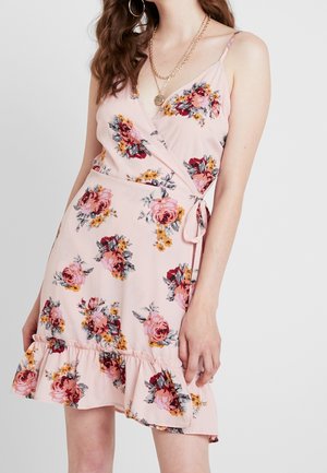 Woman wearing a pink floral wrap dress with spaghetti straps, a ruffled hem, and layered gold necklaces.