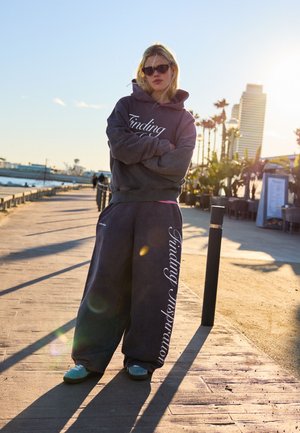 Mlada oseba, ki nosi sončna očala in preveliko temno kapuco ter hlače z napisom "Iskanje navdiha", stoji na sončni promenadi ob obali.