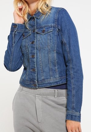 Kobieta w dopasowanej niebieskiej kurtce dżinsowej z zapięciami na guziki i szarych spodniach, dotykająca blond włosów przy twarzy.