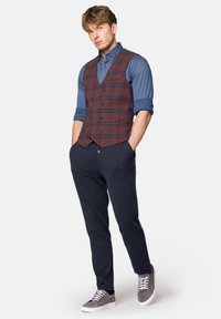 Brązowa i niebieska kamizelka w kratę na niebieskiej koszuli, w zestawieniu z granatowymi joggerami i szarymi sneakersami, tworząc warstwowy, casualowy strój.