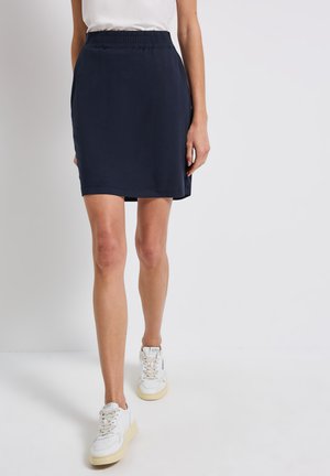 Jupe bleu marine à taille élastique, longueur genoux avec une texture lisse et une coupe décontractée. Portée avec des baskets blanches pour le contraste.