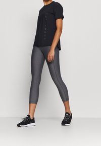 Musta urheilupaita, jossa on logotyyli, yhdistetty harmaisiin lyhyisiin leggingsseihin. Mallilla on jalassaan mustat juoksukengät, joissa on sileä pinta.