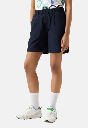 Person iført marineblå shorts med elastisk talje, hvide ankelsokker, hvide sneakers med grønne snørebånd og en mønstret skjorte, stående med den ene hånd afslappet.