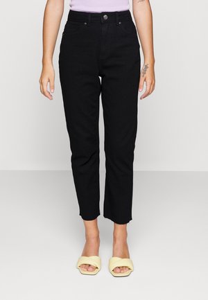 Vrouw draagt zwarte high-waist jeans met rauwe zoom, gecombineerd met lichtgele open teen slippers, staand tegen een effen lichte achtergrond.