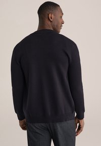 Tamsiai mėlynas megztinis su gofruota apvaliame iškirpte ir ilgomis rankovėmis. Minkšta megzta medžiaga su lygiu paviršiumi. Subtilios šoninės įpjovos apačioje.