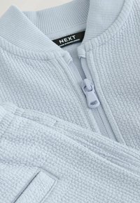 Jasnoniebieska kurtka na zamek wykonana z teksturowanej tkaniny. Posiada prążkowany kołnierz oraz smukły zamek błyskawiczny z materiałowym uchwytem.