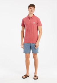 Rode polo shirt met korte mouwen en drie knopen, logo-embleem op de borst, gecombineerd met blauwe denim korte broek en bruine slippers.