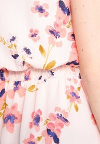 Gros plan d'une robe fleurie rose clair avec des motifs floraux bleus et jaunes et une taille élastique froncée, montrant une partie d'un bras nu.
