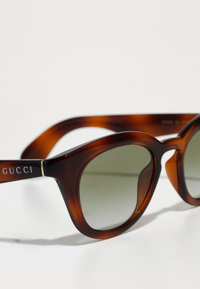 Naočale u boji kornjače s okruglim okvirom, zelenim gradijentnim staklima i metalnim detaljima. Ugravirana oznaka "GUCCI" na drškama.