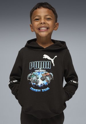 Un garçon souriant portant un hoodie noir Puma avec des camions monstres animés et le texte "Zone de Crash", les mains dans les poches, devant un fond gris uni.
