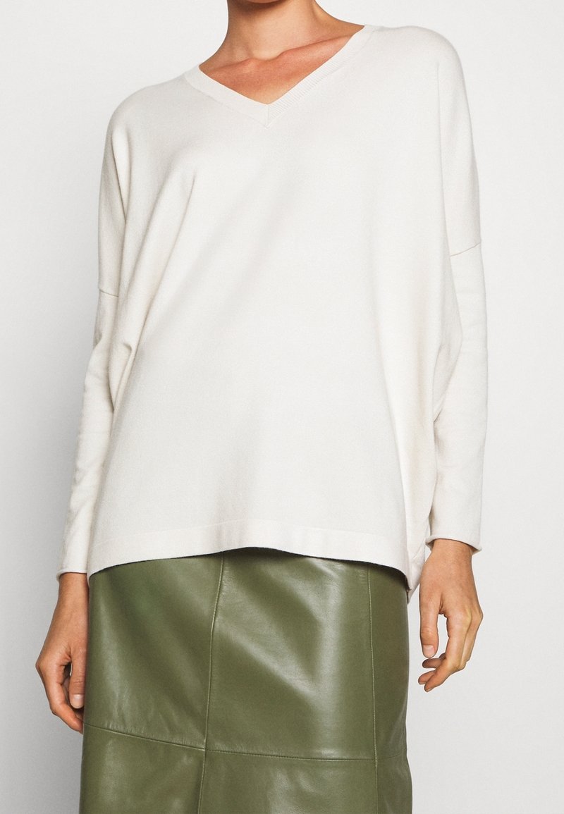 Pull ample beige clair à col en V avec manches longues, associé à une jupe en cuir vert olive taille haute. Texture lisse et design décontracté.