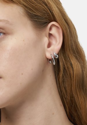 Silberfarbene Ohrringe mit einem gedrehten Design, das sanfte Kurven und glänzende Akzente aufweist. Am Ohr getragen, zeigen sie eine moderne Ästhetik.