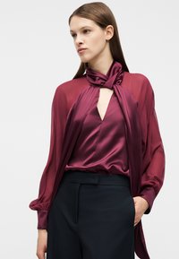 Γυναίκα που φοράει διαφανή μπορντό μπλούζα με δεμένο ψηλό γιακά και σατέν κορσάζ, συνδυασμένη με ψηλόμεση σκούρα παντελόνια, με το χέρι στην τσέπη.