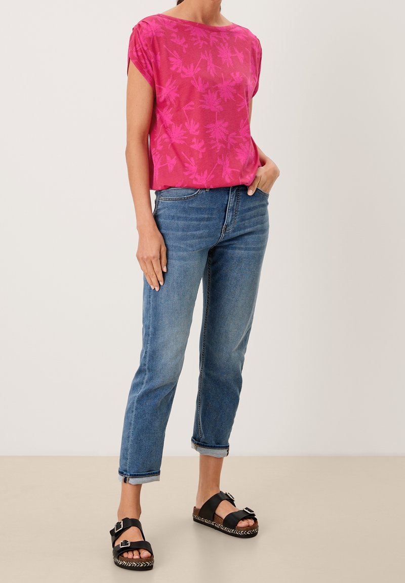 Femme portant un chemisier rose imprimé floral à manches courtes, un jean bleu retroussé et des sandales noires à double boucle, debout avec une main dans la poche.