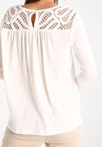 Vit långärmad topp med ett texturerat spetspanel bak, en nyckelhålöppning och draperad tyg för en avslappnad passform.