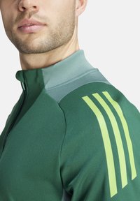 Giacca sportiva verde scuro con colletto alto e texture liscia. Presenta tre strisce sottili di un verde chiaro a contrasto sulla spalla.