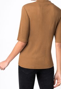 Hnědý žebrovaný top s krátkým rukávem a vysokým límcem. Materiál vypadá měkce a přiléhavě, zvýrazňuje siluetu v kontrastu s černými džíny.