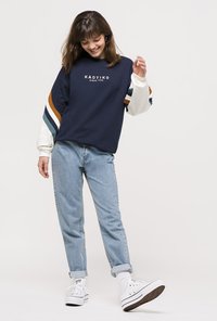 Marineblå sweatshirt med hvide og brune accentstriber på ærmerne, prydet med et logo. Lys blå denim jeans og hvide sneakers fuldender looket.