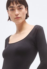 Juodas ilgomis rankovėmis viršus su plačia, kvadratine iškirpte. Pagamintas iš lygaus, prigludusio audinio, kuris tvirtai priglunda prie kūno, pabrėždamas figūrą be raštų.