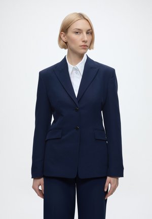 Vrouw met kort blond haar draagt een marineblauw colbert over een wit overhemd met kraag, staand tegen een effen witte achtergrond.
