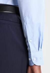 Lichtblauwe longsleeve met knoopmanchetten, gecombineerd met donkerblauwe broeken en een zwarte leren riem, waarbij de stoftextuur en pasvorm zichtbaar zijn.
