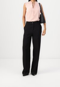Blouse sans manches rose clair avec col à volants, associée à un pantalon noir large et sur mesure, avec un bouton à l'avant et des poches.