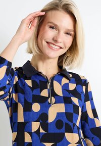 Un pulóver con cuello de media cremallera, que presenta una tela texturizada con patrones geométricos en azul, negro y beige. Mangas cortas y diseño acanalado.
