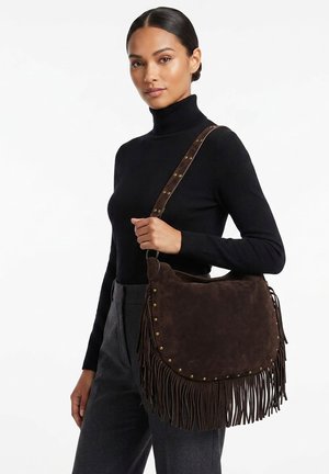 Donna con dolcevita nero e pantaloni grigi che tiene una borsa in camoscio marrone con frange e tracolla borchiata sulla spalla.